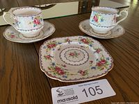 Two Royal Albert Petit Point bone china teacups on their matching saucers and a square plate with floral designs and gold trim on a wooden surface.