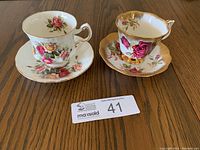 Two bone china teacups with matching saucers shown side by side on wood surface. One set has red roses and gold accents, the other lighter roses with pink and yellow colors and gold rims.