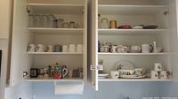 Open kitchen cabinet with four shelves containing assorted ceramic mugs, a colorful teapot, and clear glassware