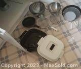 Overview of frying pan with white lid, two colanders, and wire mesh items arranged on a tiled surface.