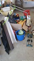 Overview of various garden tools, fabric rolls, watering cans, and seed/fertilizer spreader arranged on the floor.