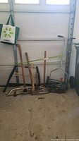 Wide view of garden tools including manual push mower, axe handles, saw, and cultivator arranged against garage wall.