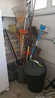 Photo of multiple wooden and metal gardening tools including rakes, shovels, hoe, broom, and blue-handled cultivator, arranged inside and next to two large black waste bins.
