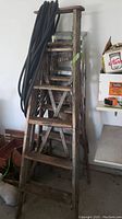 Front and side view of taller wooden ladder showing weathered steps and frame, garden hose draped over top