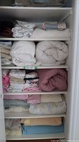 Multiple shelves with folded and rolled linens including towels, tablecloths, sheet sets, duvets, quilts, and mattress pads, mostly in neutral and pastel tones.