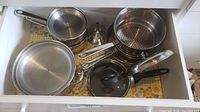 Drawer with various stainless steel Manor House pots, pans, and lids, including a strainer and steamer basket