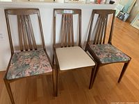 Front view of three Mid-Century Modern teak dining chairs showing two floral and one beige upholstered seats.