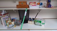 Photo shows an office supply assortment including greeting cards, gift bags, pens, a black metal mesh waste basket, a white personal paper shredder in box, and two wooden boxes on a white shelf.