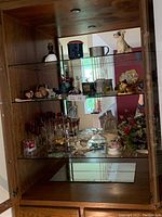 Wide view of glass cabinet with assorted figurines on upper shelves including animal figurines and fine china pieces, crystal stemware on lower shelves.