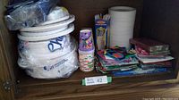 Photo showing stacks of white Chinet disposable plates, colorful paper cups with cartoon graphics, multiple colorful napkins and clear drinking straws all stored inside a wooden hutch cubby.
