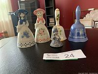 Wide photo showing six porcelain bells arranged on a table: two girl figurine bells in front, three handbells in the back row, and one smaller blue girl figurine bell placed between them.