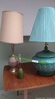 Photo of 3 lamps on wooden table: large teal ceramic lamp with pleated shade, beige ceramic lamp with beige shade, small green ceramic lamp without shade.