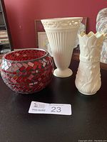 Three vases on dark table: tall cream porcelain vase by Skye McGhie, smaller cream Belleek bone china vase with floral embossing and crown rim, and red glass mosaic candle bowl.