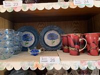 Shelf with 8 clear glass dessert bowls with bluebell flower detail stacked, two blue floral painted glass plates behind, and two groups of 5 red mugs with strawberry design on right.