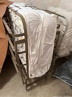 Photo showing two folded rollaway beds with metal frames and mattresses, stored upright, needing cleaning.