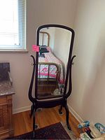 Full front view of the cheval floor mirror showing the glass and dark wood frame, standing on hardwood floor next to a wall and window.