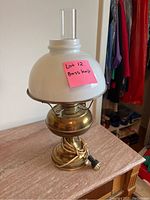 Full view of the brass lamp on a surface with electric cord coiled at base, white glass shade, and clear glass chimney.