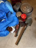 Photo showing blue REI sleeping bag, metal pots and pans with lid, camping shovel, hatchet with sheath, metal canteen, and stove fuel canister placed on the floor.