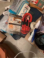 Photo showing jumper cables, windshield cover in packaging, black rubber floor mats in background, flashlight, wastebasket, and some packaged items