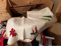 Beige tree skirt with embroidered holiday motifs including candles, poinsettia, and Christmas trees