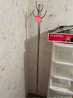 Photo of a tall metal coat rack with six hooks, standing against a floral wallpaper corner near a plastic drawer unit and a box.
