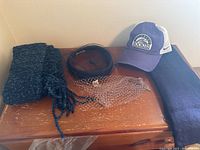 Dark blue knit scarf, black headband, fine netted veil accessory, purple and white Colorado Rockies baseball cap on wood surface