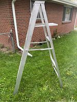 Aluminum step ladder folded, standing on grass near brick house.