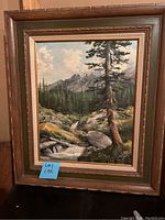 Full view of framed landscape painting showing mountains, river, trees, and large rocks under a partly cloudy sky.