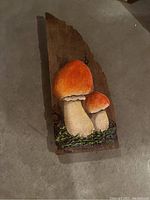 Wooden panel featuring two prominently orange-capped mushrooms painted in 3D detail on a natural wood slab with moss base, signed Fisher.