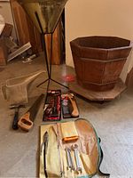Wooden planter, metal funnel with tripod, metal watering can, hand saw, rasp file, wrenches in canvas roll, metal prying tool arranged on floor