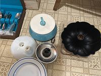 Photo showing kitchen items including blue and white ceramic pot, silver metal strainer/juicer, black bundt cake pan, and white ceramic lid with two holes.