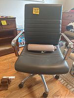 Front view of rolling office chair with black padded seat and backrest, chrome armrests, and base. Chair has a small pillow on seat.