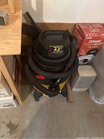 Photo of black and yellow shop vacuum with hose and nozzle wrapped on top, on concrete floor near other items.