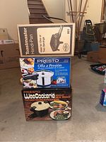 Stacked boxed appliances in basement setting: Toastmaster hand pan on top, Presto pressure cooker in the middle, Presto Weecookerie whole meal maker on bottom.