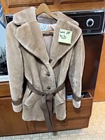 Tan faux fur coat hanging on wooden hanger with belt tied, showing wide lapel collar and accents.