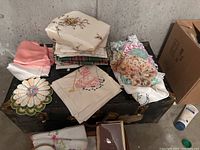 Top view of vintage trunk with stacked lace doilies, embroidered linens, silk pillowcases, and folded fabrics