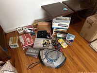 Various boxed and loose electronics including RCA boombox CD player, cables, and boxes of accessories on wooden floor near wall.