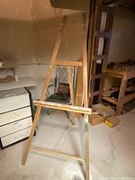 Wooden art easel in a workshop/storage area with various tools and furniture around it.