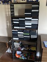 Full view of wooden bookcase with five shelves. Upper shelves contain black and white DVD or video game cases stacked horizontally. Lower shelves contain magazines, papers, and miscellaneous items including a baseball cap and small tools.