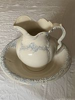 Photo showing a cream ceramic jug and bowl with gray ornamental floral and scroll detailing, jug has ornate handle and pouring spout, bowl has raised decorative rim.