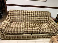 Front view of three-seat couch with beige and brown plaid upholstery, showing three cushions and padded armrests on tiled floor.