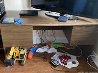 Photo showing a wooden desk with dark brown finish. The desk holds a large TV on top, with various cords underneath and toys scattered near the bottom front. The desk has an open space underneath, supported by two side panels.