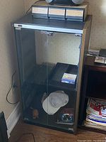 Full frontal view of the black stereo stand showcasing two glass front doors, two wooden drawers, and storage shelves with a hat and some tapes inside.