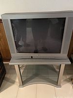 Silver CRT TV on a silver TV stand with glass shelves, located on tiled floor.