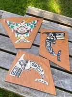 Three hand-painted wooden panels on a wooden table outdoors showing colorful bird and black and white sea creature designs in traditional Northwest Coast style.