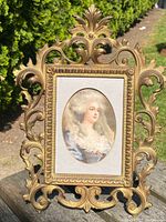 Front view of ornate solid bronze picture frame with painted oval portrait of historical woman holding as dentails of frame and portrait.