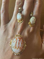 Photo showing the cameo brooch or pendant and matching earrings worn on fingers for scale and detail view of the carved classical figure cameos with gold-tone floral accents