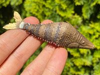 Close-up of metal articulated fish showing textured body, head, and tail fin held in fingers outside with green foliage background.