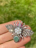 Front view of butterfly-shaped silver brooch showing pearl cluster, ruby eyes, emerald abdomen and marcasite stones on wings