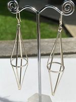 Front view of the pair of real silver geometric drop earrings on a display stand with outdoor background.